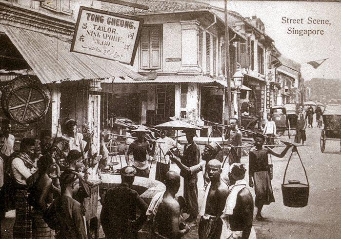 A gathering of street hawkers and others at the junction of South Bridge Road and Cross Street in Chinatown, Singapore. Cross Street was a busy street in the city popular for its many street shows and shops. Although in Chinatown, the district between Cross Street and the nearby Chulia Street was home to a substantial Indian community. The image  reflects the multi-ethnic nature of Singapore society. The focus of this photograph is the butchery located at junction of South Bridge Road and Cross Street, next to Tong Cheong Tailor.  An excited crowd of buyers as well as transport providers seems to have gathered ahead of a festive occasion, possibly Hari Raya or Muharram, which would be a day for the consumption of meat, possibly as mutton briyani (fragrant rice cooked with mutton).  The butchery gave Cross Street its Tamil name "katthikadai sadakku" (Butchery Junction).