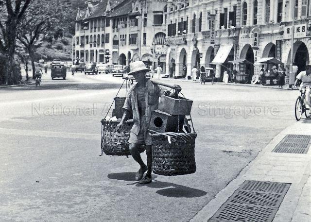 A street hawker at Bras Basah road in the 1950s. Amber Mansions located at the junction of Penang Road and Orchard Road is in the background.