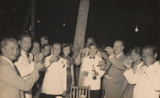 Group photograph of guests in white dinner jackets drinking a toast under the coconut trees. Chia Boon Leong is standing third from the right holding a bottle in his hand.<br />