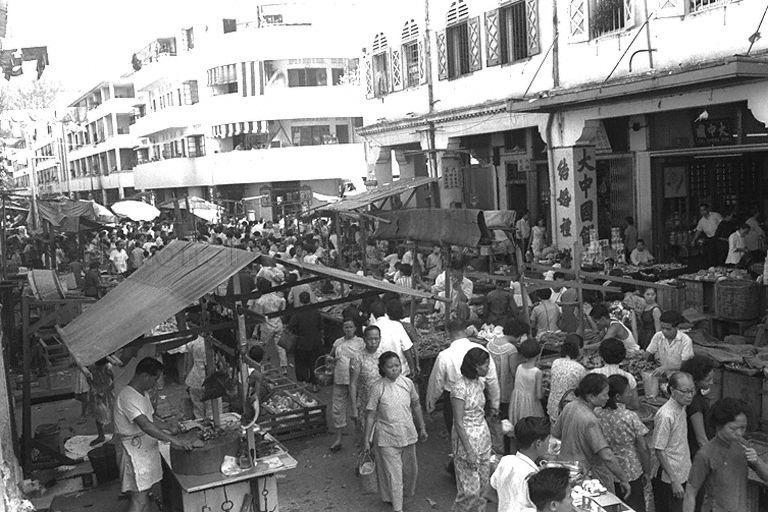 Picture of a wet market scene at Sago Street in Chinatown. The row of shophouses on the right is still around. Tai Chong Kok was at Sago Street. The pre-war Singapore Improvement Trust (S.I.T.) flats built by the British Colonial Government and available for rent only is at the top left corner of the picture.  A Chinese provision shop selling biscuits and confectionery is on the ground floor of an older part of a Chinatown building.  A butcher stall faces a row of vegetable stalls (picture).
