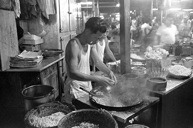 A "char kway teow" (stir-fry flat rice noodles) stall at night market. On the cabinet behind the hawker is a stack of opeh leaf (Areca catechu) which are used as wrappers for takeaway.