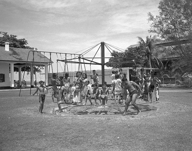 Children enjoying ride on the merry-go-round in Katong Park, during outing organised by the Joo Chiat Social Welfare Centre. Behind them is the block of changing rooms (white building with ventilation grooves) for the Katong Park bathing pagar (behind building on the left).
