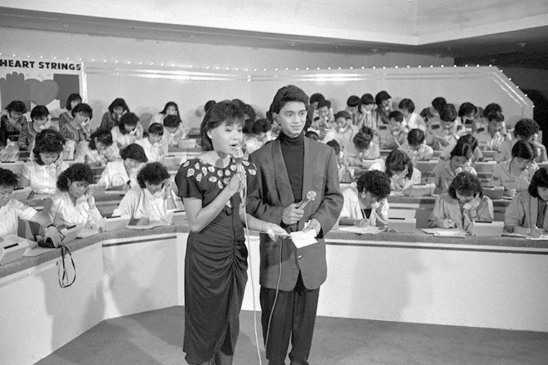 Comperes Leslie Pillay (right) and Christina Tan (left) hosting the live Community Chest's dial-a-donation "Heartstrings" charity show at Kallang Theatre, attended by President and Mrs Wee Kim Wee. In the background, volunteers from various organisations, including Ministry of Defence and Singapore Airlines, are taking calls from the public.