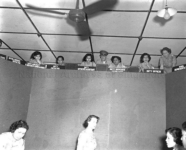General Officer Commanding of Singapore Base District Major-General A G O'Carroll Scott (centre, above) watching a squad of women volunteers at work in the controlling-reporting operation room