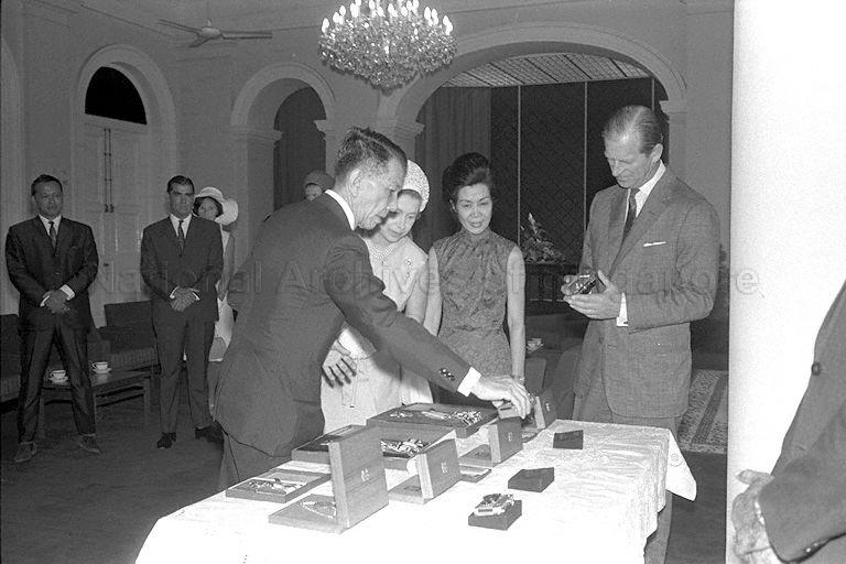 President Benjamin Sheares presenting gifts to Queen Elizabeth II, Duke of Edinburgh Prince Philip and Princess Anne, at the Istana State Room. Gifts on display include the Order of Temasek for both the Queen and Duke, the Distinguished Service Order and jade pendant for Princess Anne, three gold-plated Rollei cameras, and three gold signet rings. Mrs Sheares (second from right), Minister for Social Affairs Othman bin Wok (background, left) and British High Commissioner to Singapore Sir Sam Falle are also present.