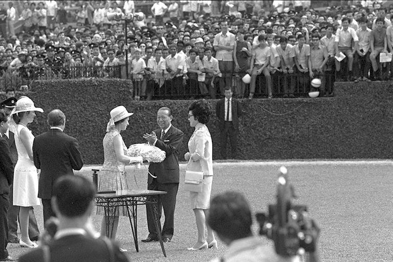 Queen Elizabeth II receiving a bouquet of flowers from wife of Chairman of Singapore Turf Club Mrs Runme Shaw, after presenting prizes of the Queen Elizabeth II Cup. Looking on are (from right to left) Runme Shaw, club secretary D R H Brown (back to camera) and Princess Anne.