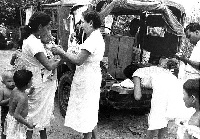 View of Dr Lam Sian Lian (bending down near vehicle) and mobile immunisation team carrying out work at a kampong. Dr Lam retired as Chief Executive Officer of Health Promotion Board in September 2005.