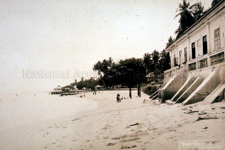 View of beach along Tanjong Katong, on east coast of Singapore. On the right are seaside residences with steps leading down to the beach. The Foreshores Act required seaside houses be built at least 50 feet (or 15 metres) from shoreline. In the background are swimming enclosures and outhouses built on piles over the water. Outhouses, a common feature of seaside houses, varied from simple open pavilions to fully furnished chalets used for picnics and parties. Many of these were demolished during World War II and remaining ones all but disappeared when coastal land was reclaimed in the 1970s.