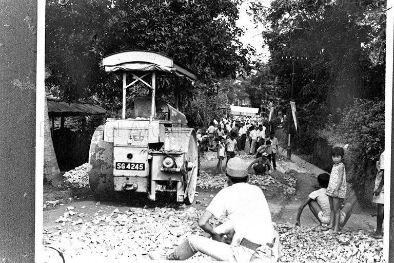 Road metalling works using crushed hardcore. An example of "gotong royong" (mutual assistance) involving kampong residents of various ages. Such "communal construction" initiatives were popular in the 1960s and 1970s of Singapore. Visible on the left is a roadworks roller bearing the old Public Works Department (PWD) logo.