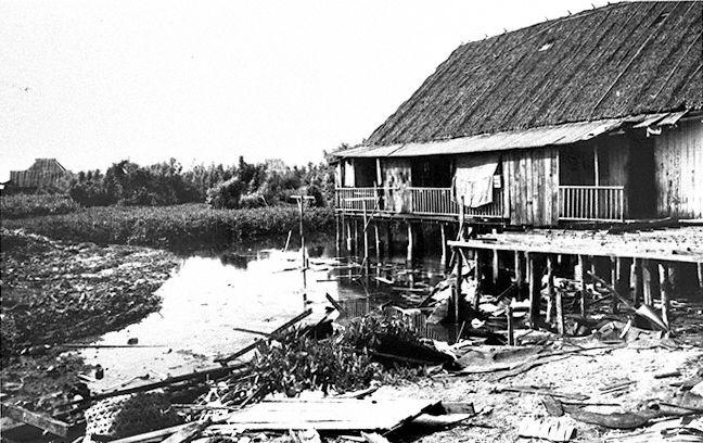 View of attap hut on stilts beside a heavily silted up waterbody, with large amounts of rubbish on the bank