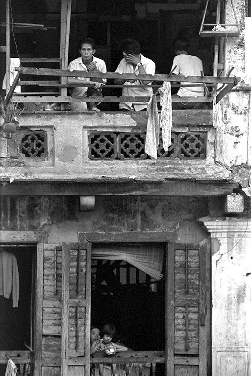 Upper levels of a shophouse in the Chinatown area. Men engage in conversation on one level while children have a meal a level below them. An urn for religious worship hangs outside the window. A wooden box (right) based on the pulley method is used to haul food bought from street hawkers to the upper floors.