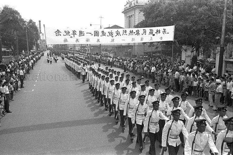 National Day Parade 1966 at the Padang - Guard of Honour contingent from the Singapore Infantry Regiment marching along the street below banner put up by residents of Hong Lim in celebration of the first National Day Parade after independence