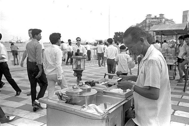 National Day Parade 1966 at the Padang - Hawker selling "kueh tutu", a local snack, along Queen Elizabeth Walk at the Esplanade where a crowd has gathered for the fireworks display. According to the family of Tan Bee Hwa, daughter of Tan Eng Huat who created the original kueh tutu, the man selling "kueh tutu" in the photo is Tan Kim Poh 陳金宝, younger cousin of Tan Eng Huat.
