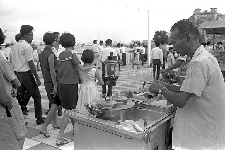 National Day Parade 1966 at the Padang - Hawker selling "kueh tutu", a local snack, along Queen Elizabeth Walk at the Esplanade where a crowd has gathered for the fireworks display. According to the family of Tan Bee Hwa, daughter of Tan Eng Huat who created the original kueh tutu, the man selling "kueh tutu" in the photo is Tan Kim Poh 陳金宝, younger cousin of Tan Eng Huat.