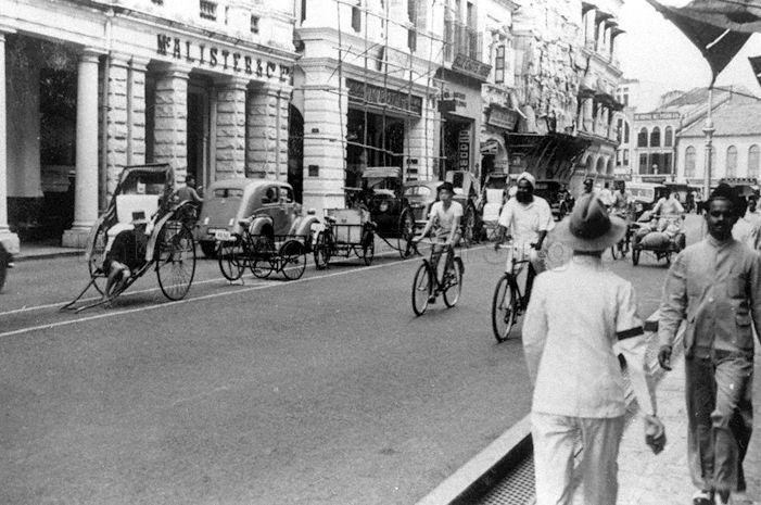 Street scene at Battery Road, Singapore, featuring pedestrians and various modes of transport at the time: motor cars, trishaws, rickshaws and bicycles
