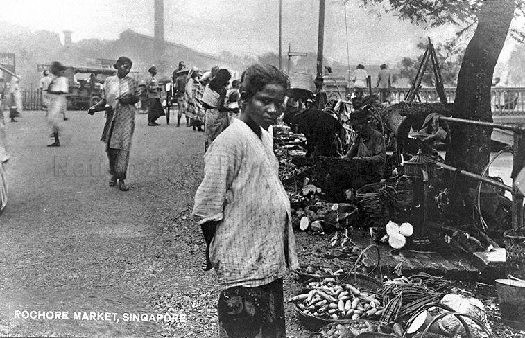 Indian lady at Rochore market, Singapore. In the background are two chimneys belonging to Straits Ice Works (nearer) and Municipal Council Incinerator further away at Kelantan Lane. The bridge on right built over Rochore Canal connects Weld Road to Arab Street on the other side. Colonial action against cotton processing in India left many weavers without a livelihood. Many from the regions of Kadayanallur and Tenkasi arrived as families in Singapore and Malaya in the early 1900s. While the men went to work in the Tanjong Pagar area, the women operated in open markets, providing small food items, especially blended spices.