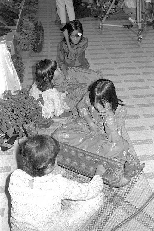 Two pairs of children playing traditional games. One pair is playing "five stones" (background) and the other pair playing "congkak" (foreground) during opening of folk arts and craft exhibition at Singapore Conference Hall. The five stones are shaped like small dumplings and usually sewn from cloth and filled with rice grains or red/green beans. Congkak are usually made of wood shaped like a boat with holes filled with marbles, cowrie shells or seeds (such as Tamarind or Red Saga seeds).