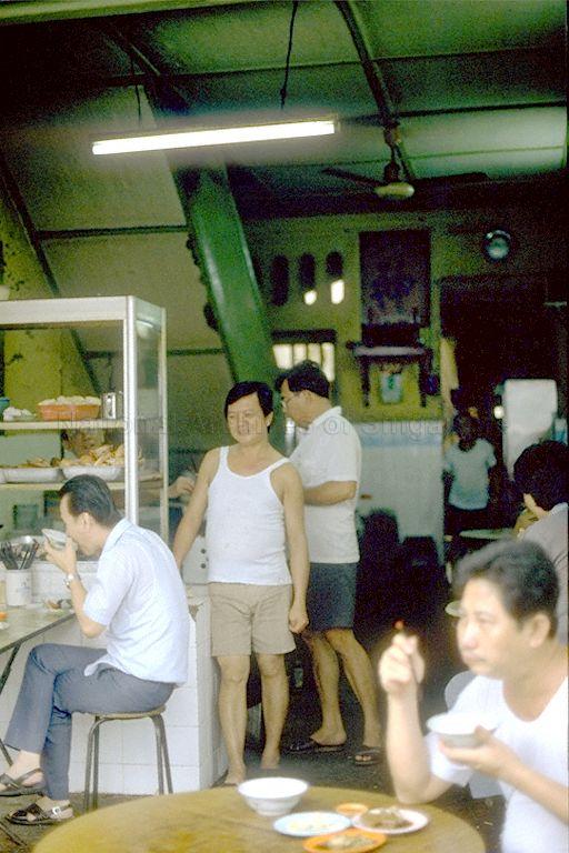 The man in the white singlet facing the camera was a kitchen assistant nicknamed 'Guni Ter' (Teochew for 'suckling pig'). His actual name is unknown. The man in glasses behind the display counter on the left of the photo is the late Ng Seow Kee, Ng Wei Chian's eldest paternal uncle, who ran the bak kut teh and Teochew porridge business in the shophouse pictured here.<br /><br />In the doorway in the background is Ng Wei Chian's second paternal uncle, Ng Seow Khern.