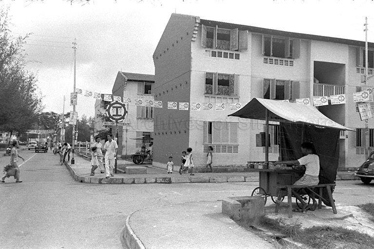 Banners of Singapore People's Alliance at Buller Terrace during the Legislative Assembly General Election 1959. The push cart stall seen here was a means of survival for Ho Khek Choon's family who resided at Block 56, 20B Buller Terrace. The cost price of one packet of cigarettes of 10 sticks was 35 cents and they were unpacked and sold at 5 cents a stick. Brands of cigarettes include Players, 555, Lucky Strike etc.