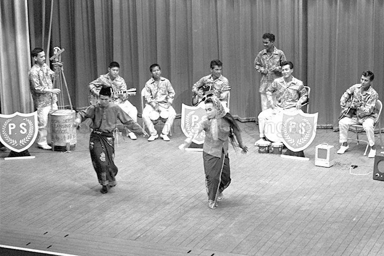 A Malay song and dance number put up by prisoners from Changi prison, Outram Road prison and Pulau Senang penal island, in aid of the Singapore Aftercare Association. In the background are seven musicians while two dancers dressed in traditional male and female Malay costumes perform a Malay dance.