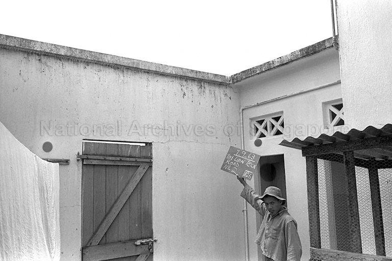 Pre-condition survey photograph No.: 126 for House No. 2 along Tay Lian Teck Road taken on 7 September 1963. A construction worker holding a written chalk board standing in front of a house wall that has visible crack lines that extend horizontally from the top of the door frame. Pre-condition survey was a documentation process in which existing faulty features of a house would be recorded by photographs before physical construction works for the new sewer line commenced in the vicinity of the house. This photographs would be referred to for legal purpose should dispute occurs to ascertain whether defects were already inherent in the building prior to construction of the new sewer line.