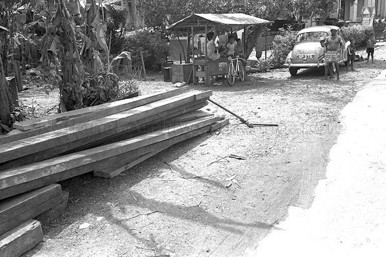 Drainage and sewerage works by Public Works Department (PWD). The PWD worker is holding a chalk board in front of a house at No. 96 Devonshire Road. Beside him is likely a Morris Minor car with license plate number SP579. A bicycle with a license plate attached beneath its seat is seen in front of a roadside hawker stall where three boys are resting on. Licenses were required for bicycles in the early days of Singapore.
