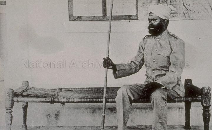 Pavement-based occupations -- A Sikh watchman in uniform holding a stick and sitting on his charpoy or bedstead