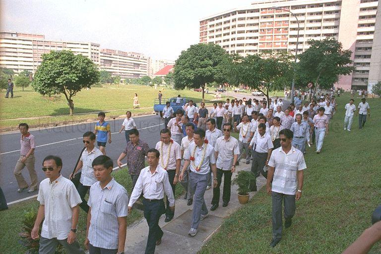 Minister for Trade and Industry and Second Minister for Defence Brigadier-General Lee Hsien Loong and the ministerial delegation touring Nee Soon Central constitency. From left (garlanded) are Member of Parliament (MP) for Nee Soon Central Ng Pock Too, BG Lee, MP for Punggol Ng Kah Ting, Minister of State for Foreign Affairs and National Development Peter Sung, (mostly hidden), and MP for Hong Kah Group Representative Constituency (GRC) John Chen.