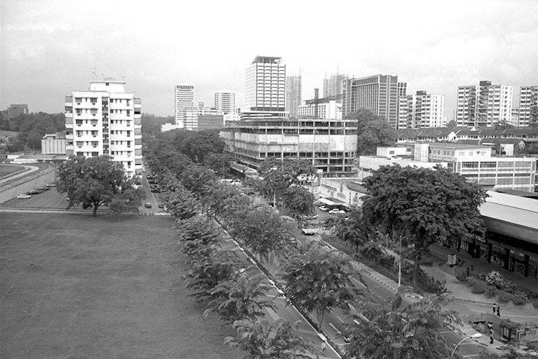 View of Orchard Road from Mandarin Hotel, showing (from right middle-ground) Fitzpatrick's Supermarket, Lucky Plaza (a 30-storey shopping and residential building) in construction, and Shaw Centre behind it. The field in the left foreground is where Ngee Ann City would be built. Hyatt Regency Singapore (the tall building in darker shade with curved top edges), opened in 1971, can be seen in the background on the right.