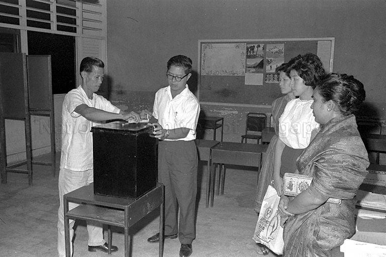 Election officials sealing a ballot box while three ladies, who are probably witnesses, look on during Legislative Assembly General Election 1963. Tables and chairs in the room and a notice board on the wall behind suggested that this was a classroom in a school was used as a polling station.