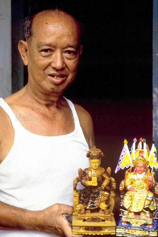Ng Tian Sang of "Say Tian Hng Buddha Shop" at 6 Gemmill Lane holding wooden carvings of Chinese deities