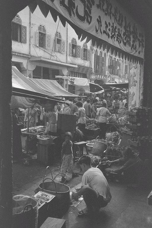 View of wet market along Sago Street in Chinatown from five-foot-way outside shop selling the sweet and sticky "nian gao" or Chinese new year cake (in circular containers held by elderly woman on the right). Since this dessert used to be sold only during the Chinese or Lunar New Year season, this photo would have been taken between January and February that year. Diagonally opposite is the famous Tai Chong Kok Chinese confectionery.