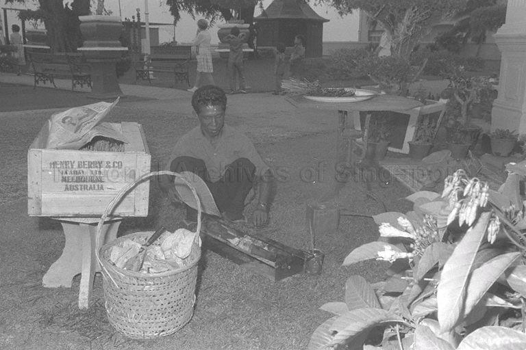 "Satay" seller fanning charcoal in portable grill to cook for birthday celebration of Madam Tan Teck Neo at Mandalay Villa, No. 29 Amber Road. Madam Tan, born 18 December 1877, was the second wife of Straits-born Chinese businessman and philanthropist Lee Choon Guan. In the background are children and guests in the garden of this grand bungalow which was built in 1902 and well known for parties thrown by Madam Tan that brought the Chinese and British communities together in those days when they seldom mixed.