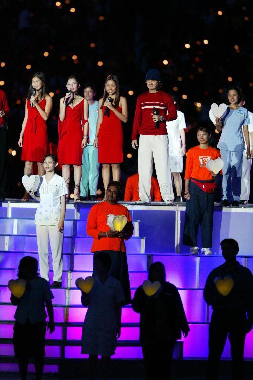 National Day Parade 2003 Preview at National Stadium -- View of emcees and celebrities (in red, from left) Wong Li Lin, Sharon Au, Jean Danker and Gurmit Singh singing theme song "Through Your Eyes", inspired by the SARS episode. They are joined by MediaCorp artistes, including May Phua (next level below, in orange), and 240 health-care workers, each holding a glowing paper heart.