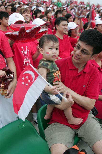 National Day Parade 2008 at Marina Bay - Toddler with stick-on face tattoo and holding a Singapore flag amongst cheerful spectators dressed in National flag colours of red and white