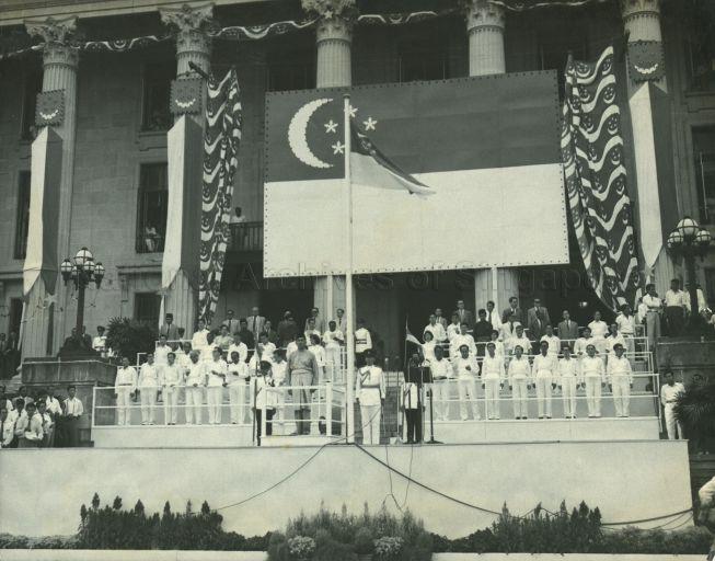 Singapore was proclaimed a self-governing state on 3 June 1959. This was celebrated with National Loyalty Week which was held between 3 to 10 December 1959 and commenced with the installation of Yusof bin Ishak as the first Malayan-born Yang di-Pertuan Negara (Head of State). This photograph shows the newly-installed Yang Di-Pertuan Negara and the Cabinet and Legislative Assembly Members at the City Hall steps during the launch of National Loyalty Week. The new state flag is prominently displayed.