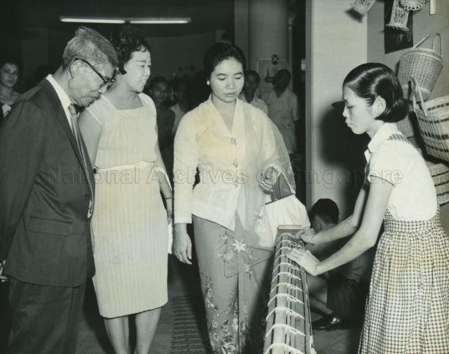 Puan Noor Aishah watching a blind girl making rotan chicks at the Sale of Work by the Singapore Council of Social Welfare at Victoria Memorial Hall. On the left is philanthropist Lee Kong Chian.