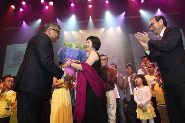 Chief Justice Sundaresh Menon (on the left) is presenting Justice Lai Siu Chiu with a bouquet of flowers. Looking on are Chairman of the Yellow Ribbon Fund Phillip Tan (on the right) and Attorney-General Steven Chong (in a maroon shirt). They are on stage with the performers during the Singapore Academy of Law Charity Concert. The concert is held at School of the Arts, Drama Theatre.