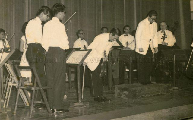 Conductor Goh Soon Tioe and members of the Goh Soon Tioe String Orchestra taking their bow after performance.Goh Soon Tioe String Orchestra at concert featuring Ma Si-Hon as soloist. Goh Soon Tioe on podium and Ma Si Hon next to him taking a bow. Photo taken approximately around 1962.