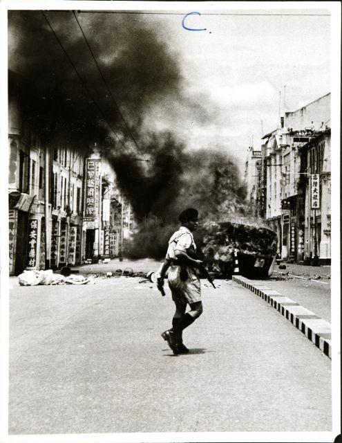 This photograph illustrates one of the incidents in the Report on Military Action taken in the Singapore Riots, 25 October to 2 November 1956. Police in the foreground  with a burning vehicle in the background.