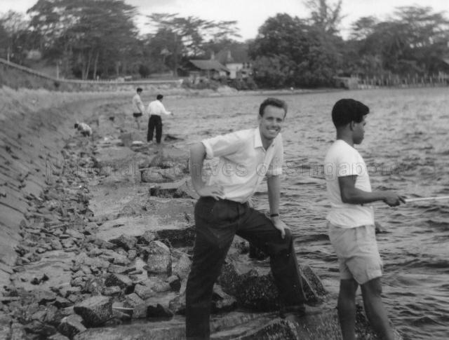 Photograph of George Phillips standing on the rocks with local fishing enthusiasts at the Singapore - Johore Causeway
