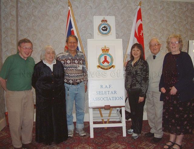 Photograph of Joan (second from left), Major Yeo Kuan Joo and his wife (centre), Arthur Davey, Rita (left) and other member posing with the Royal Air Force Seletar Association (RAFSA) poster at the RAFSA reunion